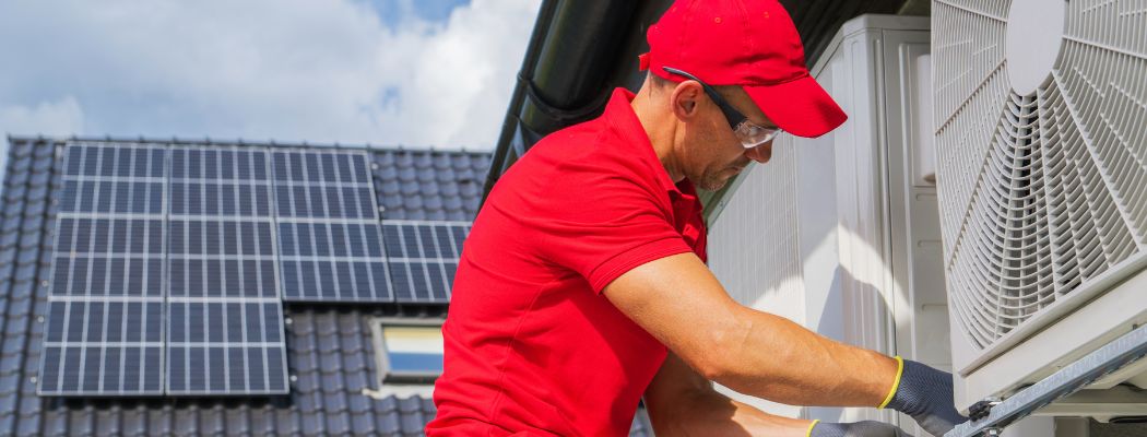 Mann mit rotem T-Shirt und Cap installiert Wärmepumpe. Im Hintergrund ein Dach mit Solarmodulen